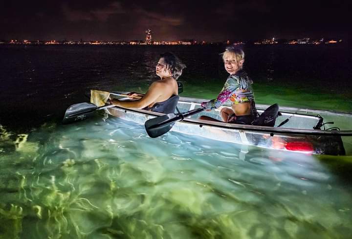 Two guests paddling a clear LED-lit glass bottom kayak over glowing shallow water at night, with the Sarasota skyline behind them