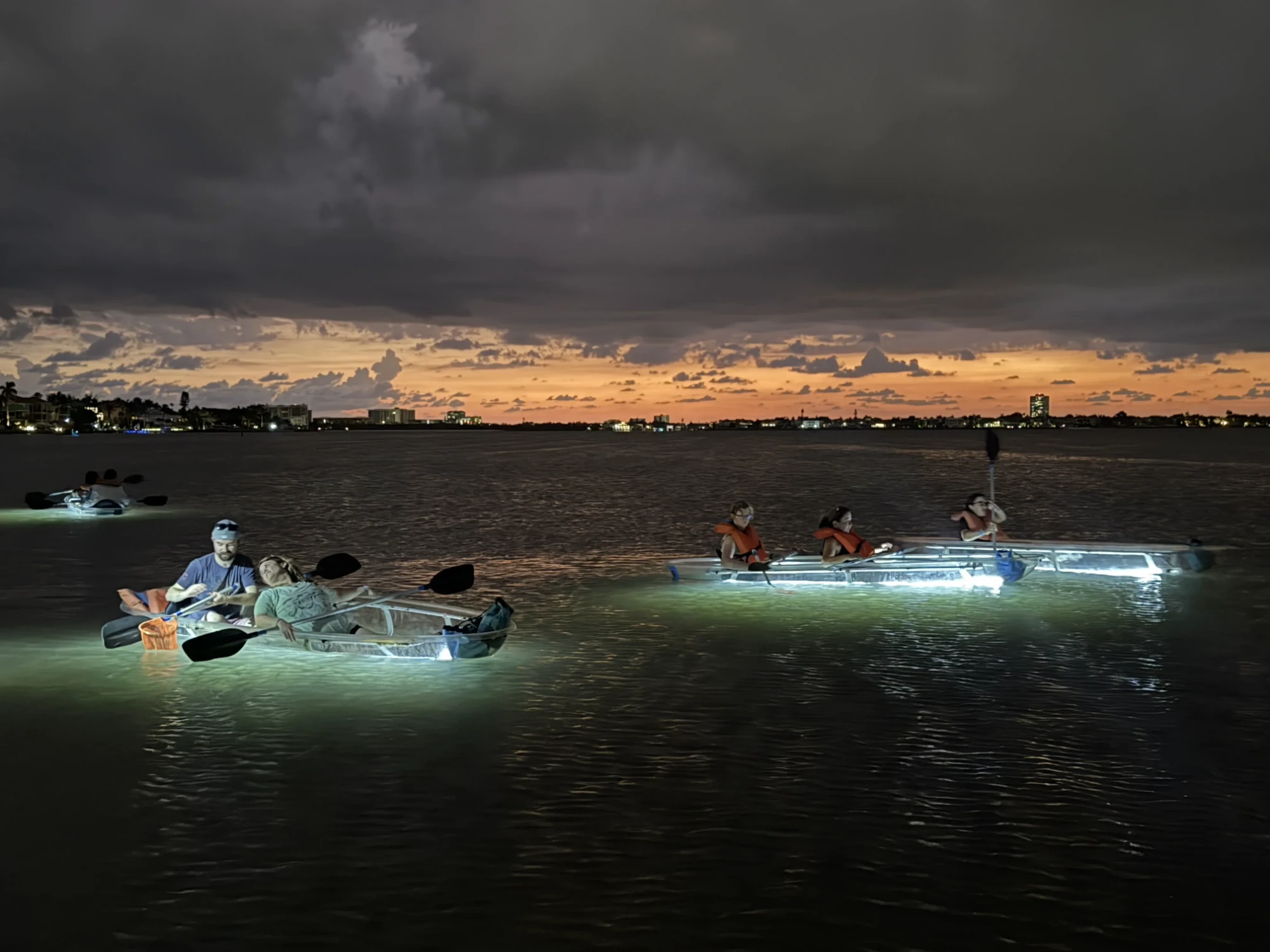Three LED-lit clear kayaks glowing on the shallow flats at dusk, with the Sarasota skyline and an orange sunset on the horizon